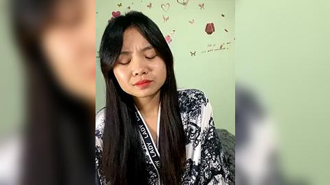 Media: Video of a young Asian woman with long black hair, wearing a black and white patterned robe, sitting in a green room with butterfly and floral wallpaper.