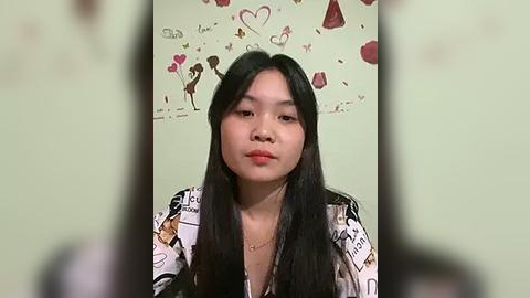 Media: A video of an East Asian woman with long black hair, wearing a patterned top, standing against a wall adorned with heart-themed wallpaper.