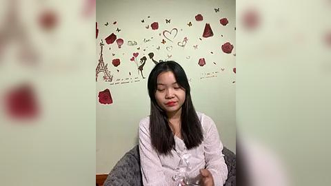 Media: Video of an East Asian woman with long black hair, wearing a white blouse, seated in a room with a Paris-themed wallpaper featuring Eiffel Tower and heart designs.