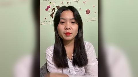 Media: Video of an East Asian woman with straight black hair, wearing a white lace top, standing against a pastel green wall adorned with heart and flower decals.