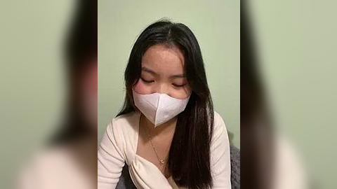 Media: Video of an Asian woman with long black hair, wearing a white face mask and a beige cardigan, against a green wall, eyes closed, expression neutral.