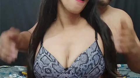 Media: Video of a woman with long black hair, fair skin, wearing a black and gray snake print bra, capturing a close-up of her cleavage. Her face is partially visible, smiling with pink lips. Background is blurred, featuring a dark curtain.