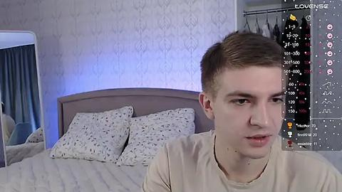 Media: Video of a young man with short brown hair, fair skin, and a neutral expression, sitting on a bed with white patterned pillows. Background features a light blue wall, a window with curtains, and a digital display showing social media statistics.