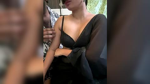 Media: Video of a woman with light skin, medium build, wearing a sheer black dress with a deep V-neckline, posing indoors. Background features a man in a patterned shirt and a partially visible floral wallpaper.