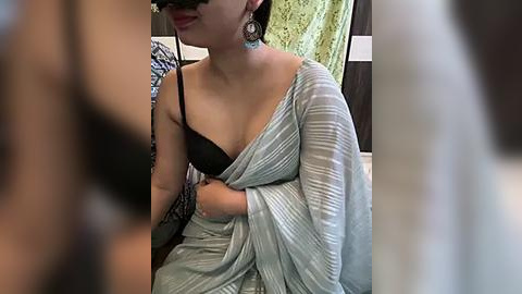 Media: A video of an Indian woman with a medium complexion, wearing a black bra under a light blue, pleated sari, sitting in a dimly lit room with a white towel and patterned fabric in the background.