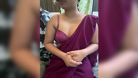 Media: Video of a light-skinned woman with curly hair, wearing a magenta sari with a lace bra, sitting indoors with patterned cushions and curtains in the background.