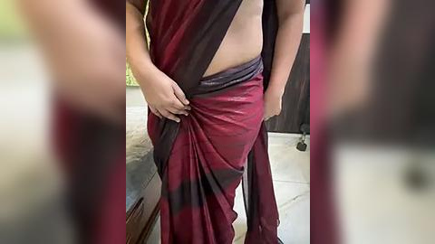 Media: A video of a person wearing a maroon and black striped sari, holding it up to reveal their midriff, in an indoor setting with a blurred background.