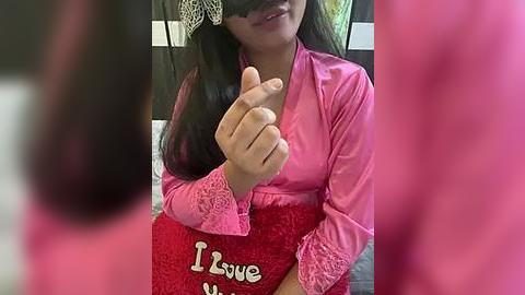 Media: Video of a smiling woman with long black hair, wearing a pink satin robe and a red \"I Love You\" pillow, indoors with blurred background.