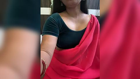 Media: Video of a South Asian woman with medium skin tone, long black hair, wearing a dark green short-sleeved blouse and a vibrant red sari, her face partially obscured by another person's arm.