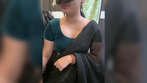 Media: Video of a woman in a teal top and black sari, wearing a blindfold, with blurred figures in the background.