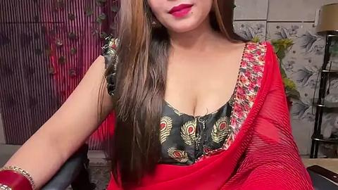 Media: Video of a South Asian woman with long brown hair, wearing a red saree with floral patterns, showcasing her ample cleavage, seated indoors with a maroon curtain and floral wallpaper in the background.