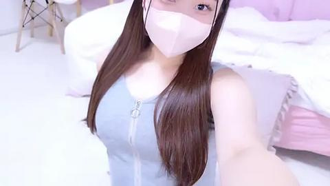 Media: Video of an East Asian woman with long brown hair, wearing a pink face mask and a light blue zip-up top, lying on a white bed with pink pillows and a gold metal frame in the background.