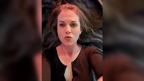 Media: A close-up video of a middle-aged Caucasian woman with fair skin and short brown hair, lying on a dark bed. She wears a black top, looking serious and slightly pensive.