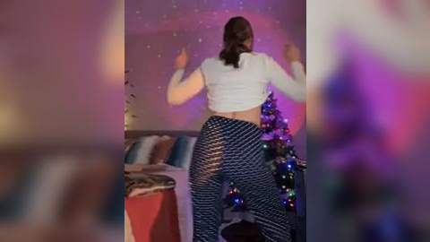 Media: Video of a woman in a white crop top and patterned pants, dancing energetically in a living room with a lit Christmas tree, festive lights, and a red couch.