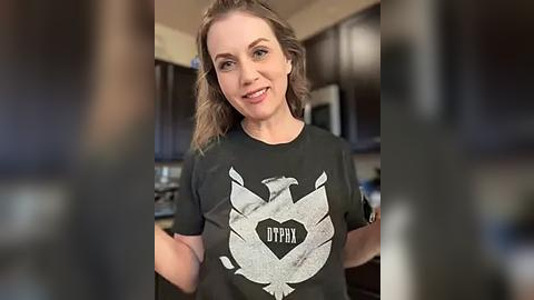 Media: Video of a smiling, fair-skinned woman with wavy brown hair, wearing a black T-shirt with a large, stylized white eagle emblem and \"DYPTA\" text. Background shows a modern kitchen with dark cabinets and stainless steel appliances.