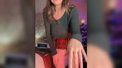 Media: Video of a woman with light skin and wavy blonde hair, wearing a green cardigan over a red plaid skirt, reaching out in a cozy, dimly lit room with a Christmas tree in the background.