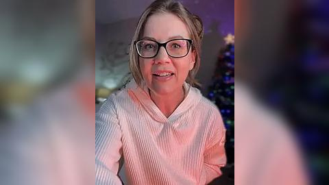 Media: A video of a smiling woman with glasses, wearing a white ribbed sweater, standing indoors with a blurred Christmas tree in the background.