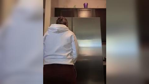 Media: Video of a woman with braided hair, wearing a gray hoodie and maroon sweatpants, standing in a kitchen, facing a stainless steel refrigerator.