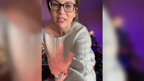 Media: Video of a middle-aged woman with glasses, wearing a textured, light-colored sweater, smiling, with a blurred, festive background featuring a Christmas tree and purple lights.