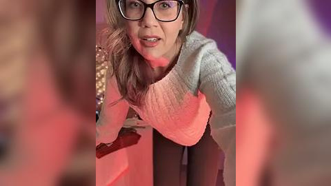 Media: Video of a woman with glasses, wearing a light sweater and black pants, bending over a table, blurred background with red and purple lights.