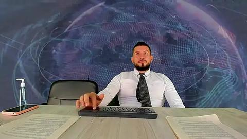 Media: Video of a bearded, middle-aged man in a white dress shirt and black tie, gesturing with his right hand at a computer screen displaying a complex, abstract, blue and purple holographic image on a light-colored wooden desk.