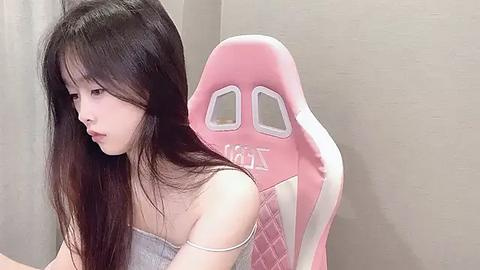 Media: A video of an East Asian woman with long, straight black hair, wearing a light gray off-shoulder top, seated in a pink gaming chair with white accents. She appears to be deep in thought, with a neutral expression. The background is a plain beige wall.