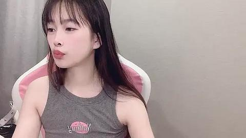 Media: Video of a young Asian woman with long, straight black hair, wearing a gray sleeveless tank top with a pink heart graphic, sitting in a white and pink gaming chair against a beige wall.