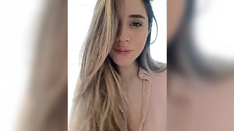 Media: Video of a young woman with long, blonde hair partially covering her face, wearing a light pink blouse, indoors with a blurred background.