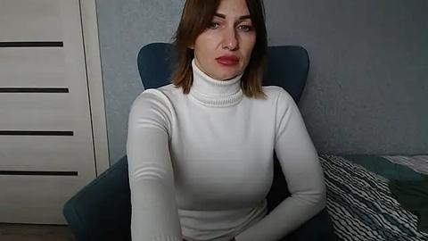 Media: Video of a fair-skinned woman with shoulder-length brown hair, wearing a white turtleneck, sitting in a teal armchair against a blue textured wall, with a striped pillow on the left.