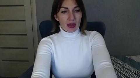 Media: Video of a young woman with light skin and straight, shoulder-length brown hair, wearing a white turtleneck sweater. She has a neutral expression and slightly open mouth. Background shows a blue chair and a zebra-patterned blanket.