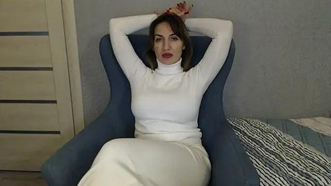 Media: Video of a light-skinned woman with brown hair, wearing a white turtleneck and white pants, sitting in a blue chair, arms raised, in a room with a striped bedspread and beige walls.