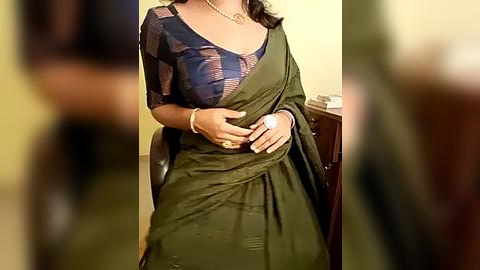 Media: Video of a woman in an olive-green saree with a sheer blue blouse, seated on a chair, wearing a silver necklace and a watch. Background includes a blurred person and a wooden desk.