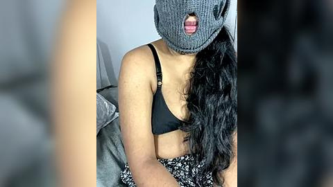 Media: Video of a woman with medium skin tone, wearing a gray knitted beanie with a cartoon face, black bra, and black and white patterned pants, sitting on a bed.