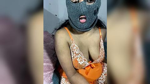 Media: A video of a woman with medium skin tone wearing an orange lace lingerie set, a grey knit beanie with eye holes, and a surprised expression.