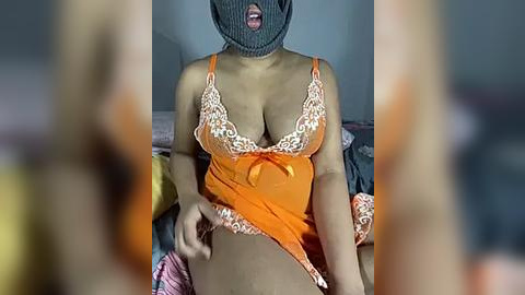 Media: A video shows a woman with medium brown skin wearing an orange camisole with lace trim, her breasts partially visible, and a black face mask. She sits on a bed with blurred background.