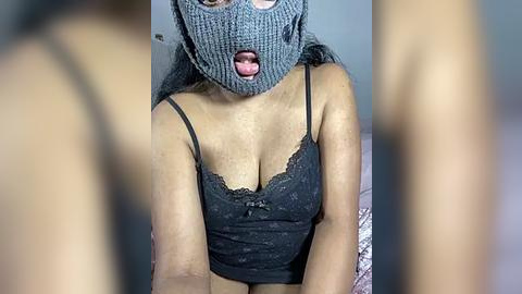 Media: Video of a light-skinned woman with medium-sized breasts wearing a gray knitted mask, black lace bra, and a black tank top, sitting on a bed, blurred background.