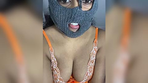 Media: Video of a woman with medium skin tone, wearing a gray knit mask and orange lace bra, her large breasts prominently displayed. Background is blurred.