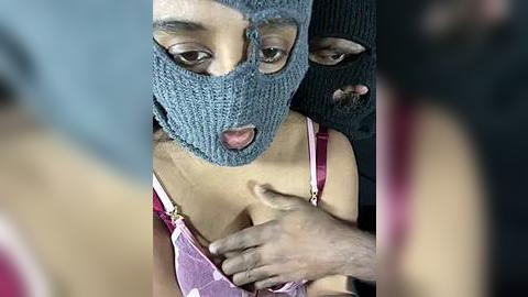 Media: Video of a woman in a gray balaclava, pink bra, and a man's hand on her chest, blurred background.