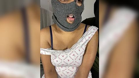 Media: Video of a woman wearing a grey knitted mask, exposing her face, and a white floral dress, indoors.