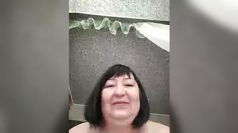 Media: A video of an older woman with short black hair, fair skin, and a slight smile, standing in a bathroom with a textured gray shower curtain and a white towel hanging on the wall.