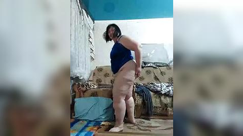 Media: A video of a plus-size woman in a blue tank top and no pants, standing in a cluttered, dimly lit room with a patterned couch, a messy floor, and a blue blanket.