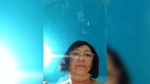 Media: Video of a middle-aged woman with short black hair, wearing glasses, against a bright blue background. The image is slightly blurry.