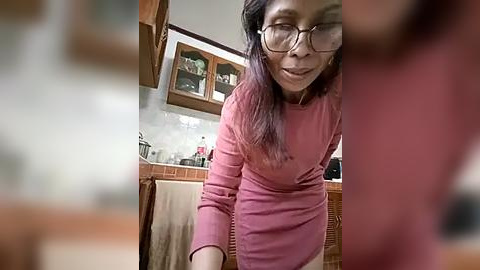 Media: Video of a smiling Asian woman with long, dark hair, wearing glasses and a pink dress, standing in a cozy kitchen with wooden cabinets, utensils, and a hanging towel.