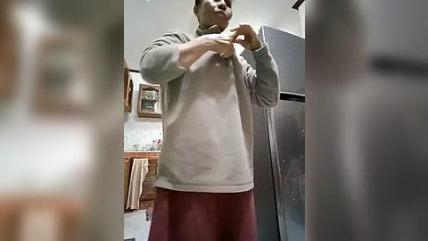 Media: Video of a person wearing a gray sweater and maroon pants adjusting a metal object in a modern kitchen with stainless steel appliances, wooden cabinets, and framed art on the wall.