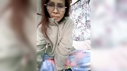 Media: Video of a middle-aged Asian woman with long brown hair, wearing glasses, a beige jacket, and floral-patterned pants, sitting on a bed with a floral-patterned sheet.