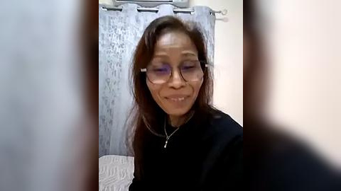 Media: Video of a smiling, middle-aged Black woman with long brown hair, wearing glasses, a black top, and a silver necklace, standing in a dimly lit bathroom with a gray curtain.