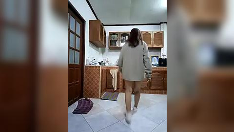 Media: A video of a woman in a gray sweater and black underwear walking away from a kitchen with wooden cabinets and white walls.