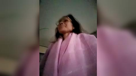 Media: A blurry video of an elderly Asian woman with long dark hair, wearing a pink bathrobe, standing in a dimly lit bathroom with a white towel in the foreground.