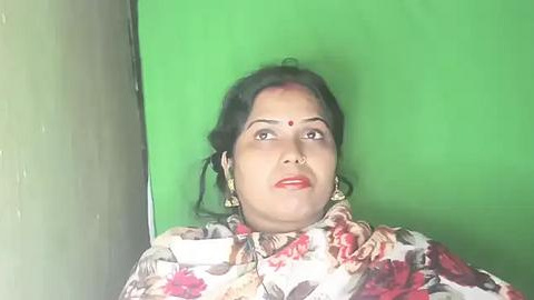 Media: Video of a woman with medium skin tone and black hair, wearing a floral blouse, against a green background, applying makeup.