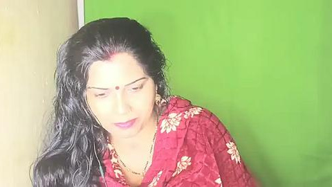 Media: Video of a South Asian woman with long, wavy black hair, wearing a red floral saree, a bindi, and a gold necklace, against a green backdrop.